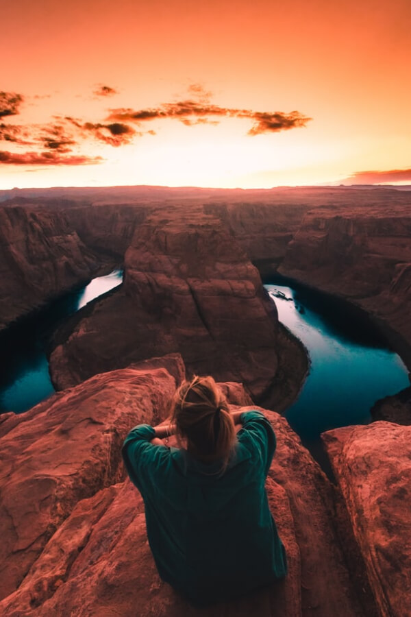 Person sitting on rocky ledge overlooking Horseshoe Bend at sunset, with vibrant orange sky and winding river below, reflecting lifestyle themes in 'Barefoot Blonde' blog post.