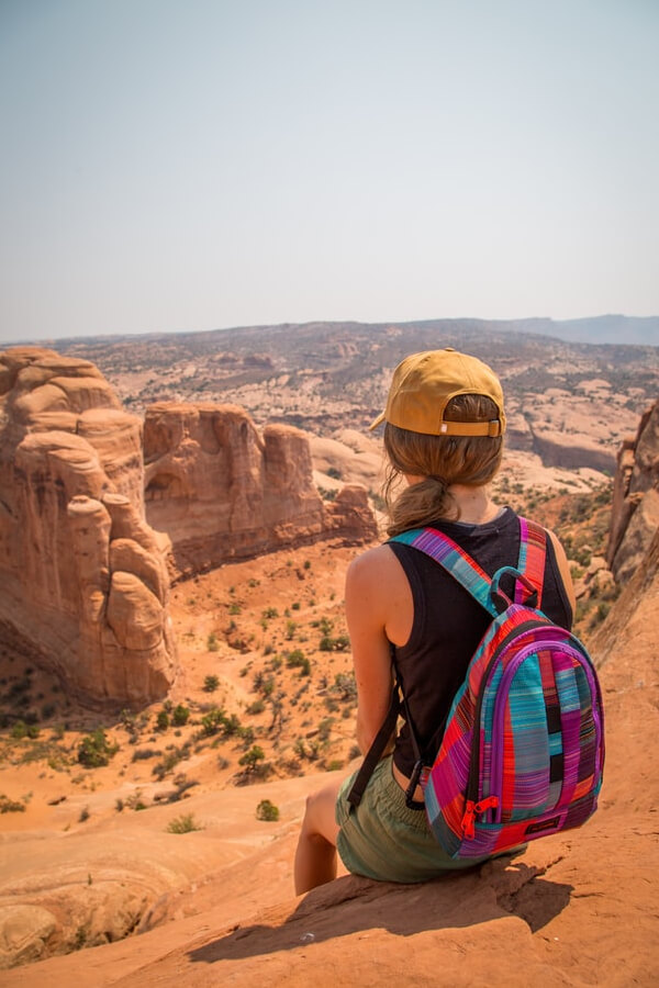 Person with a colorful backpack sitting on a rocky ledge, overlooking a vast desert landscape, embodying adventure and exploration.